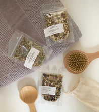 Load image into Gallery viewer, Three botanical bath tea sample pouches on a white surface with a dry brush, gray towel, and wood spoon.