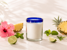 Load image into Gallery viewer, Sunset Margarita soy candle in a handcrafted Mexican artisan recycled glass with a cobalt blue rim, surrounded by pineapple, lime, mint, and hibiscus on a soft white background.