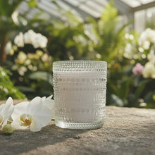 Load image into Gallery viewer, Cattleya and Ylang Ylang soy candle in clear hobnail glass jar on a stone surface with white flowers and greenery in the background.