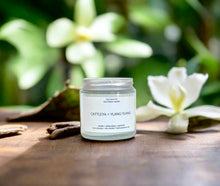 Load image into Gallery viewer, Cattleya + Ylang Ylang candle on a wooden surface with a background of white flowers and greenery.