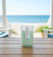 Load image into Gallery viewer, A 12 oz hand-poured coconut and soy candle in a green-tinted recycled glass jar, Black Sea scent, on a table with a beach view in the background.