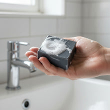 Load image into Gallery viewer, Hand holding a black charcoal soap bar with a soft-focus bathroom background.