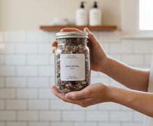 Load image into Gallery viewer, Hand holding Root Ritual botanical bath tea jar with a subway tile bathroom background.
