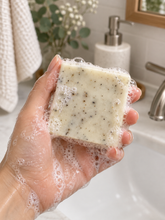 Load image into Gallery viewer, Close-up of a wet hand holding a creamy white, botanical-speckled bar of soap covered in bubbles over a bathroom sink, with a towel, greenery, and soap dispenser softly blurred in the background.