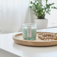 Load image into Gallery viewer, A 10 0z coconut and soy candle in a green-tinted recycled glass jar, Seven Seas scent, on a wooden tray with a blurred plant in the background. Hand-poured in Wilmington, North Carolina.