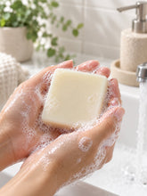 Load image into Gallery viewer, Close-up of wet, lathered hands holding a smooth, creamy white bar of soap over a running bathroom sink, with greenery and a neutral soap dispenser softly blurred in the background.