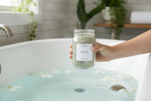 Load image into Gallery viewer, Hand holding a jar of Quiet Tide bath salt above a white bathtub filled with pale blue water, floating white flowers, and a serene spa-like setting.