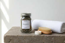 Load image into Gallery viewer, Herbal Recovery bath tea in a glass jar on a stone top with a dry brush, folded white towel and tealight.