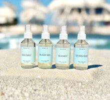 Load image into Gallery viewer, Four Sea Collection room spray bottles—Sea Salt, Black Sea, Sea Mist, and Seven Sea—displayed on sand with water and blurred boats in the background.