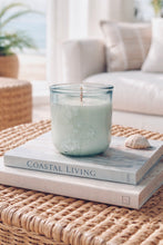Load image into Gallery viewer, Seafoam green scented candle in embossed floral glass jar on stacked books, styled on a woven coffee table in a bright coastal beach house living room.