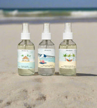 Load image into Gallery viewer, Three Vacation Collection room spray bottles—Island Escape, Wrightsville Breeze, and Kisses By The Sun—displayed on sand with a blue ocean wave in the background.