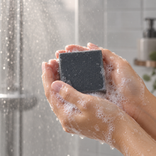 Load image into Gallery viewer, Close-up of hands lathering a dark charcoal-colored bar of soap under running shower water, surrounded by bubbles in a softly lit, neutral-toned bathroom.
