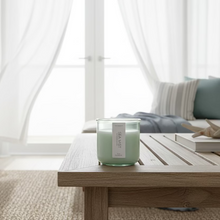 Load image into Gallery viewer, A 10 oz coconut and soy candle in a green-tinted recycled glass jar, Sea Mist scent, on a light natural brown coffee table in a coastal living room. Hand-poured in Wilmington, North Carolina.