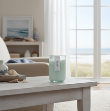 Load image into Gallery viewer, A 12 oz hand-poured coconut and soy candle in a green-tinted recycled glass jar, Sea Mist scent, on a whitewashed end table in a beach cottage.