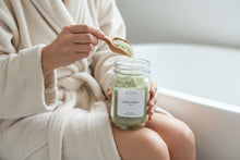 Load image into Gallery viewer, Person in a bathrobe scooping Ocean Purity bath salt from a glass jar with a wooden spoon beside a bathtub in a calm, spa-like setting.