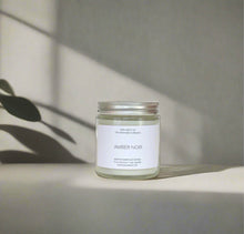 Load image into Gallery viewer, AMBER NOIR candle styled on a light concrete surface, green leaf in the back with a shadow falling over
