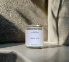 Load image into Gallery viewer, AMBER NOIR candle on a smooth stone surface with a shadow falling on the texture stone wall.