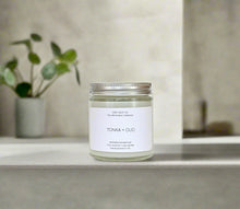 Load image into Gallery viewer, Tonka + Oud candle on a raw texture surface with a plant in a modern, urban space