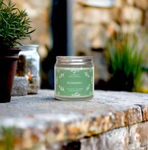 Load image into Gallery viewer, Fresh rosemary candle on a stone surface, with herb pots and a stone wall in the back