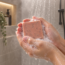 Load image into Gallery viewer, Close-up of hands lathering a pink, speckled bar of soap under running shower water, with soft neutral tiles, an amber bottle, and greenery in the background.
