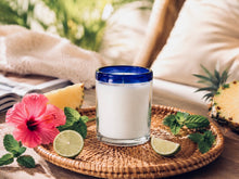 Load image into Gallery viewer, Sunset Margarita soy candle from SIRA NEST NC in a handcrafted recycled Mexican artisan glass with a cobalt blue rim, styled on a woven tray with pineapple, lime, mint, and hibiscus in a bright, coastal-inspired home.
