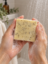 Load image into Gallery viewer, Close-up of wet, lathered hands holding a pale yellow bar of soap speckled with dark botanical particles under running shower water, with greenery and an amber pump bottle softly blurred in the background.
