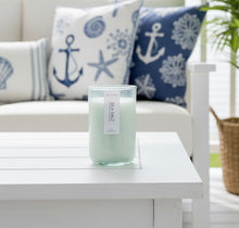 Load image into Gallery viewer, A 12 oz hand-poured coconut and soy candle in a green-tinted recycled jar, Sea Salt scent, on a table with decorative pillows featuring anchors and starfish in the background.
