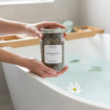 Load image into Gallery viewer, Hand holding a jar of Herbal Recovery bath tea above a white bathtub filled with pale blue water, floating white flowers, and serene spa-like setting.
