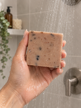 Load image into Gallery viewer, A hand holding a rectangular bar of speckled dusty rose pink soap under running shower water in a tiled bathroom setting.
