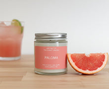 Load image into Gallery viewer, A grapefruit wedge and a Paloma cocktail are placed on a light wooden surface next to a Paloma candle.
