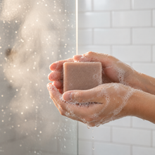 Load image into Gallery viewer, A close-up of hands holding a rectangular bar of soap in front of a shower glass, with soap foam covering the hands and water droplets falling in a bright bathroom setting.
