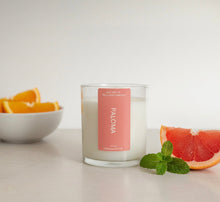 Load image into Gallery viewer, Clear tumbler jar candle placed on a light stone surface next to a bowl of oranges and a slice of grapefruit, evoking a refreshing citrus and tequila-inspired scent.
