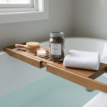 Load image into Gallery viewer, Root Ritual botanical bath tea in a glass jar on a bathtub caddy with a dry brush, tealight and folded white towel over a tub filled with pale blue water.
