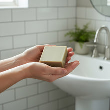 Load image into Gallery viewer, Hands holding an olive green soap bar with a soft-focus bathroom background.
