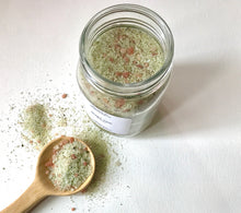 Load image into Gallery viewer, Open jar of Quiet Tide bath soak with a wooden scoop filled with green and pink mineral salts on a white background.

