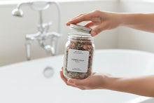 Load image into Gallery viewer, Hand holding Evening Calm bath tea while opening the jar lid above a white bathtub.
