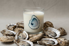 Load image into Gallery viewer, Candle in a glass jar with oyster shell design surrounded by real oysters on a neutral background
