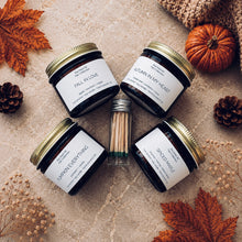 Load image into Gallery viewer, Four amber glass fall candles with gold lids—Fall in Love, Autumn in My Heart, Pumpkin Everything, and Spiced Maple—arranged around a vial of matches on a natural stone surface with autumn leaves, pinecones, dried florals, and a small pumpkin.
