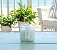 Load image into Gallery viewer, A 10 oz coconut and soy candle in a green-tinted recycled jar, Sea Salt scent, on a table with a patio background. Hand-poured in Wilmington, North Carolina.
